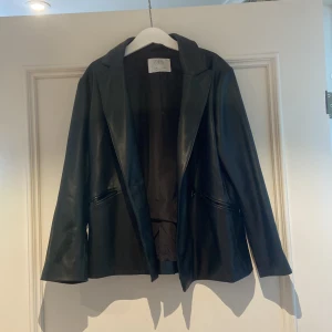 Svart skinnkavaj från Zara - Svart kavaj i skinnimitation från Zara med klassisk krage och snygga detaljer som tre knappar vid ärmslutet och fickor med dragkedja. Jackan har en stilren och rak passform med långärmade ärmar, perfekt för dig som gillar en edgy look.