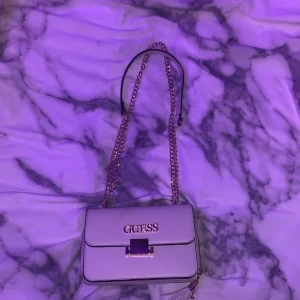 Vit axelväska från Guess med kedja - Snygg vit axelväska från Guess med guldfärgad kedja och logga framtill. Väskan har ett stilrent, fyrkantigt format och flera fack med dragkedja. Materialet är syntet och den har en dekorativ tofs på sidan. Perfekt för att hålla ordning på dina grejer.