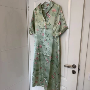 Grön blommig långklänning - Snygg långklänning i ljusgrönt satinmaterial från Lost Scene med blommigt mönster i rosa och vitt. Klänningen har korta ärmar, krage och knappar framtill. Perfekt för dig som vill ha en chill och elegant look med lite vintagevibe.