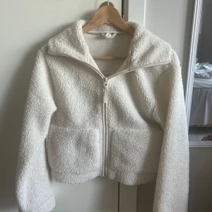 Vit teddyjacka med dragkedja, S - Mysig vit teddyjacka från H&M i storlek S. Jackan har fluffig yta, två stora fickor framtill och hel dragkedja. Perfekt för kalla dagar och har en avslappnad look med hög krage och lång ärm.