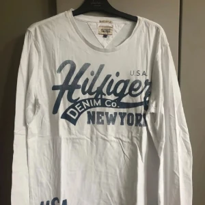 Vit långärmad Hilfiger Denim tröja - Vit långärmad tröja från Hilfiger Denim med stort blått tryck på bröstet där det står 'Hilfiger Denim Co. New York'. Rund halsringning och mjukt bomullsmaterial. Perfekt för dig som gillar streetwear och klassisk amerikansk stil.