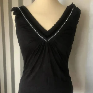 Svart topp med v-ringning och pärlor - Svart topp med snygg v-ringning som är dekorerad med pärlor längs kanten. Toppen har lätt rynkade detaljer framtill och små volanger vid axlarna. Modellen är ärmlös och har en slim passform som framhäver siluetten. Perfekt för dig som gillar stilrena detaljer.