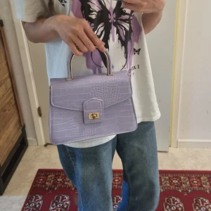 Bijou Brigitte axelremsväska - Säljer en super gullig ljuslila handväska från Bijou Brigitte i croco-imitation med guldfärgade detaljer och vridlås. Väskan har ett metallhandtag och avtagbar axelrem. Väskan har tecken på användning men har varit väl omhändertagen. Storlek 23 x 5 x 17cm