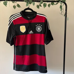 Tysklands VM-tröja 2014 Adidas - Säljer en officiell Tysklands fotbollströja från VM 2014 med breda svarta och röda ränder, broderade FIFA World Champions-märke och landslagsemblem. Adidas-logga på bröstet och vita ränder på axlarna. Tröjan är kortärmad och tillverkad i lätt polyester.