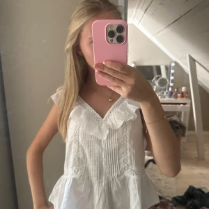 Vit blus med volang och spets - Supersöt vit blus med volang vid v-ringad hals och spetsdetaljer längs framstycket. Blusen är ärmlös och har en peplumformad nederdel som ger en chill och feminin vibe. Perfekt till jeans eller kjol för en fräsch look.