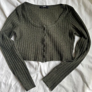 Olivgrön ribbad croppad kofta Bik Bok - Säljer en olivgrön croppad kofta från Bik Bok med ribbad struktur och långa ärmar. Koftan har en snygg rundad halsringning och knappar framtill. Perfekt att styla med höga jeans eller kjol för en chill look.