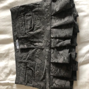 Grå minikjol med volang  - Säljer en grå minikjol med coolt ormmönster och volangdetalj nedtill. Kjolen har klassiska jeansfickor både fram och bak samt bälteshällor. S/M