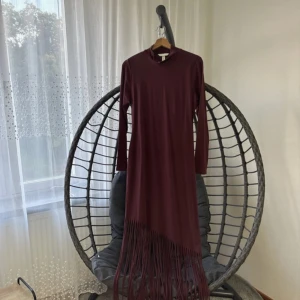 Vinröd långklänning med fransar från H&M - Säljer en vinröd långklänning från H&M i mjukt material med lång ärm och hög hals. Klänningen har coola fransdetaljer nedtill som ger extra edge. Perfekt för dig som gillar att sticka ut med stilrena detaljer och djup färg. Storlek M