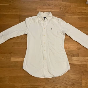 Vit skjorta från Polo Ralph Lauren XS - Vit klassisk skjorta från Polo Ralph Lauren i bomull, custom fit. Skjortan har lång ärm, knappar framtill och den ikoniska broderade loggan på bröstet. Perfekt krage med knappdetaljer och stilren look som funkar till jeans eller chinos.