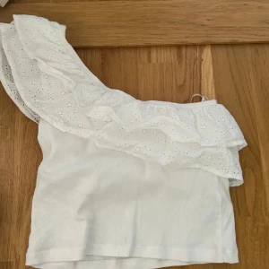 Vit oneshoulder topp med volang - Supersöt vit topp med oneshoulder-design och bred volang med broderade hålmönster. Ribbad botten och luftig känsla, perfekt för varma dagar. Volangen ger en extra touch av romantisk stil.