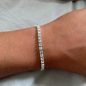 Silverfärgat tennisarmband med stenar - Stilrent tennisarmband i silverfärg med gnistrande klara stenar runt hela bandet. Armbandet har en klassisk, smal design som ger en lyxig känsla och passar perfekt för dig som gillar att sticka ut med bling. Enkel att matcha med andra smycken för en extra touch.