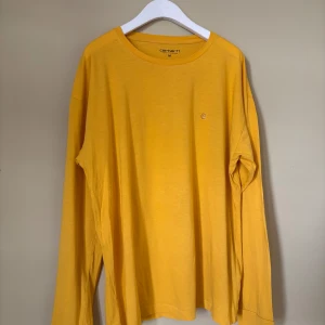 Gul långärmad tröja Carhartt M - Gul långärmad tröja från Carhartt i storlek M. Tröjan har en enkel design med rund hals och liten broderad Carhartt-logga på bröstet. Tillverkad i mjuk bomull som är skön mot huden. Perfekt för dig som gillar streetwear och vill ha en färgklick i garderoben.