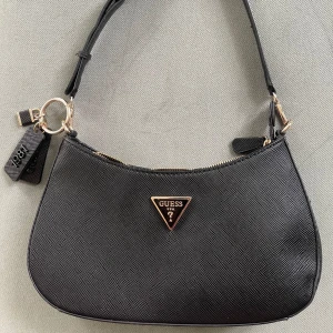 Guess Axelremsväska svart - Säljer en få gånger använd äkta Guess väska i svart. Köpt på zalando för lite över 1000 kr. Säljer då den ej kommer till användning längre. Fortfarande i toppen skick, har kvitto till denna , Mått Höjd 18 cm Längd 29 cm Vidd 7 cm