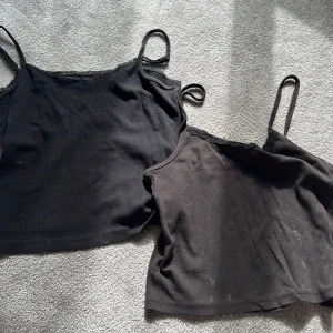 Svarta croppade linnetoppar med spets - Två svarta croppade linnetoppar från Basic U med tunna axelband och dekorativ spets längs halsringningen. Materialet är ribbad bomull som ger en skön och stretchig känsla. Perfekt för att styla lager på lager eller till jeans och kjol. Lite smutsiga och slitna men fina ändå!