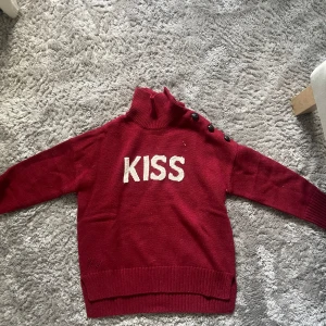 Röd stickad polotröja med KISS-tryck - Snygg mörkröd stickad polotröja med vit broderad KISS-text framtill. Tröjan har lång ärm, ribbade muddar och coola svarta knappar längs axeln. Perfekt för dig som gillar statement-plagg och vill sticka ut lite extra.