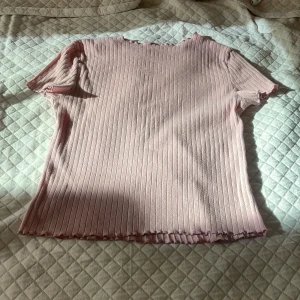 Ljusrosa ribbad topp med volangkant - Supersöt ljusrosa topp med ribbad struktur och volangkant vid hals, ärmar och nederkant. Modellen är kortärmad och har en slim passform som sitter snyggt på kroppen.