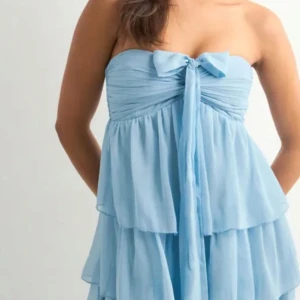 Ljusblå omlottklänning med volanger - Supersöt ljusblå kortklänning med flera lager volanger och omlottband som kan knytas både runt halsen och som rosett framtill. Klänningen är axelbandslös och har ett skirt, lätt material som ger en flowy känsla. Perfekt för sommarens alla tillfällen! Det finns en liten fläck som inte syns vid användning och en tråd som inte syns. Skriv för mer bilder.