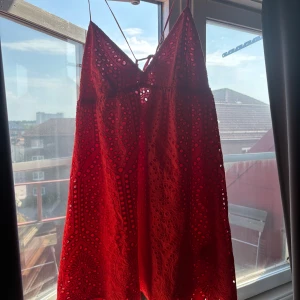 Röd kort klänning med broderi - Säljer en röd kort klänning med tunna axelband och snygg broderad spets. Klänningen har ett detaljerat mönster med blommor och cirklar, samt knytning i ryggen. Perfekt för varma dagar och har en luftig känsla tack vare bomullsmaterialet.