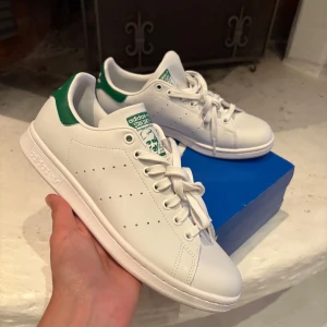Adidas Stan Smith vita sneakers strl 42 - Fräscha Adidas Stan Smith sneakers i klassisk vit färg med gröna detaljer på hälen och tungan. Skorna har rund tå, platt sula och snörning. Tillverkade i syntetmaterial med ikonisk Stan Smith-logga bak och på tungan. Perfekt för dig som gillar clean och tidlös stil.