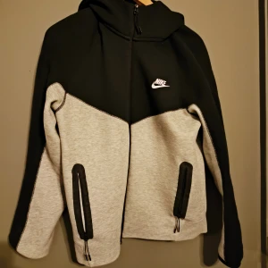 Nike tech svart och grå hoodie M - Nike tech hoodiejacka i svart och ljusgrått med dragkedja framtill och två fickor med svarta detaljer. Jackan har huva och logga på bröstet. Tillverkad i mjuk polyester, perfekt för chill eller träning. Snygg kontrast mellan färgerna och stilren design.