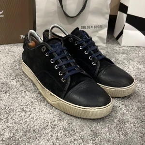 Lanvin mörkblå - Lanvin sneakers i mörkblå färg med rund tå och platt vit sula. Ovandelen är i mocka och skinn, med mörkblå snörning och metallöglor. Klassisk och stilren design som passar dig som gillar exklusiva märken och vill ha en clean look.