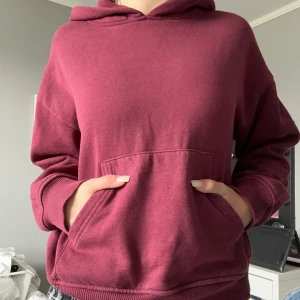 Vinröd hoodie med huva och ficka - Säljer en vinröd hoodie med stor känguruficka framtill och huva. Tröjan har en loose passform och långa ärmar med ribbade muddar. Perfekt för chill dagar och enkel att matcha med jeans eller mjukisbyxor. Materialet är mjukt och känns skönt mot huden.