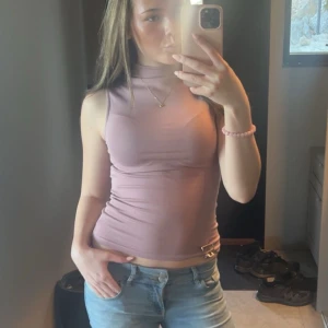 Ljuslila polotopp i stretchig bomull - Säljer en ljuslila polotopp med tight passform och ärmlös design. Toppen har en hög krage och är gjord i mjukt, stretchigt bomullsmaterial som sitter snyggt mot kroppen. Perfekt att styla med jeans eller kjol för en clean och modern look.