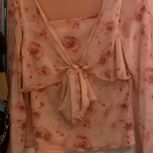 Blommig topp med knyt - Romantisk blus i ljusrosa med blommigt mönster och transparent känsla.Volangdetalj över axlarna ger extra flow. Perfekt för dig som gillar en söt och trendig look.