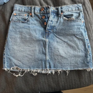 Blå jeanskjol med fransig kant - Säljer en blå jeanskjol från lager 157 med fransig, rå kant nertill och klassiska fickor både fram och bak. Kjolen har knappar framtill och bälteshällor.