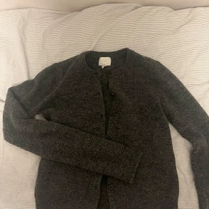Mörkgrå kofta från H&M XS - Mysig mörkgrå kofta från H&M i storlek XS. Koftan är stickad med rund halsringning och knäppning framtill. Perfekt för lager-på-lager och har långa ärmar. Enkel och stilren design som funkar till det mesta.