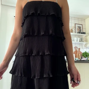 Svart volangklänning med tunna axelband - Supersöt svart klänning med flera lager volanger och tunna axelband. Plisserad struktur som ger en cool och trendig look. Perfekt för dig som gillar en lekfull och chic stil.