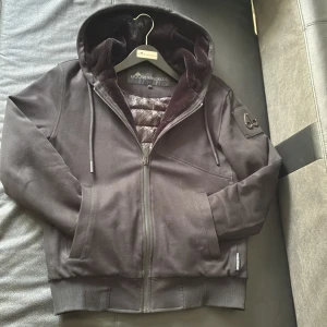 Svart hoodiejacka Moose Knuckles - Snygg svart hoodiejacka från Moose Knuckles med stor logga på ryggen och emblem på ärmen. Jackan har ett väldigt skönt material på insidan plus den är perfekt när det är kallt ute men du ej vill ha på en stor vinterjacka. Det är strolek xs men de passar S/M Perfekt för dig som vill ha en streetig och exklusiv look. Använd ett fåtal par gången men har inga skador, köpt på NK och har kvitto plus du får galgen med. Köpt för 4700 säljer för 3999, pris kan diskuteras.
