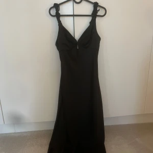 Svart långklänning med knutade axelband - Elegant svart långklänning med djup v-ringning och knutade axelband. Klänningen har en figurnära passform upptill och faller snyggt längs kroppen. 