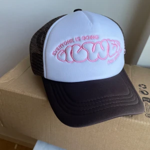 Brun och vit truckerkeps med rosa brodyr - Snygg truckerkeps med vit frontpanel och brun skärm. Kepsen har bruna mesh-sidor och baksida för extra ventilation. På framsidan finns broderad text i bubblig rosa stil och på sidan ett broderat ansikte i rosa. Justerbar baktill.