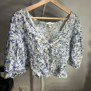 Blommig blus från H&M i linneblandning - Supersöt blus från H&M i vit och blå linneblandning. Blusen har blommigt mönster, v-ringning, knappar framtill och puffiga trekvartsärmar med resår. Perfekt för en fräsch och somrig look!