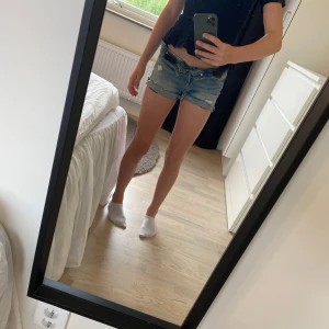 Blå jeansshorts med fransig kant - Säljer ett par klassiska blå jeansshorts med rå, fransig kant och slitna detaljer. Shortsen har fem fickor och normal passform, perfekt för varma sommardagar. De är midwaist och gjorda i denim som ger en avslappnad vibe.
