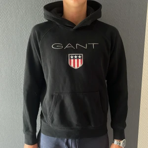 Svart hoodie från GANT med brodyr - Svart hoodie från GANT med broderad logga och sköld på bröstet. Klassisk känguruficka framtill och huva. Mjuk bomullskänsla och långärmad modell, perfekt för dig som vill vara stilig till vardags 