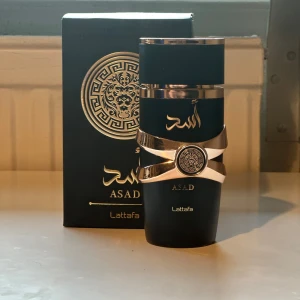 Lattafa Asad Parfym 100ml - Lattafa Asad är en parfym i en matt svart flaska med guldiga detaljer och ett lejonmotiv. Flaskan har en unik design med ett guldfärgat band och mönstrad medaljong. Förpackningen är svart med guldtryck och orientaliska inslag. Doften är kraftfull och exklusiv. Aldrig använd så helt full (100ml) skickar samma dag✅