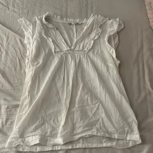 Vit blus med volang från lager 157 - Supersöt vit blus från lager 157 med volangdetaljer vid ärmar och halsringning. Blusen har v-ringning med spets och är ärmlös, perfekt för en fräsch och luftig look. Tillverkad i mjuk bomull som känns skön mot huden.