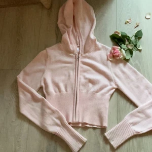 Ljusrosa hoodie från Gina Tricot - Superfin ljusrosa hoodie från Gina Tricot med dragkedja framtill. Tröjan är croppad med bred ribbad mudd i midjan och vid ärmsluten. Mjuk stickad känsla och huva för extra cozy vibe. Perfekt för dig som gillar en söt och trendig look.