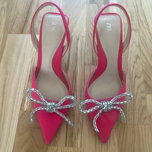 Rosa pumps med strassrosett från Zara - Nya och oanvända snygga rosa pumps från Zara med spetsig tå och dekorativa strassrosetter framtill. Skorna har öppen häl och smala remmar, samt hög klack. Perfekta för dig som vill sticka ut med en färgstark och glittrig detalj.