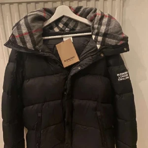 Svart dunjacka från Burberry - Snygg svart dunjacka från Burberry med klassiskt rutigt foder i huvan. Jackan har dragkedja framtill, två fickor med dragkedja och logga på ärmen. Perfekt för kalla dagar och ger en clean och lyxig vibe.