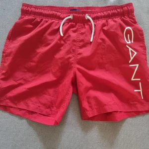 Röda badshorts från GANT med snörning - Snygga röda badshorts från GANT med vit snörning i midjan och stort vitt GANT-tryck längs ena benet. Klassisk modell med elastisk midja och snabbtorkande material, perfekta för stranden eller poolen.