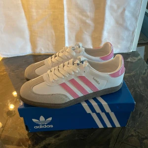 Adidas Samba vit/rosa sneakers - Snygga Adidas Samba sneakers i vitt med rosa detaljer och klassiska tre ränder på sidan. Ovandel i skinn och mocka, med grå sula och vita snören. Perfekt för dig som vill sticka ut med en fräsch färgkombo och sportig vibe.