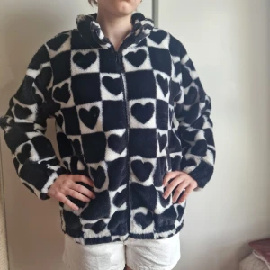 Svartvit teddyjacka med hjärtan - Fluffig teddyjacka i svart och vitt med rutigt mönster och hjärtan. Jackan har dragkedja framtill, hög krage och långa ärmar. Perfekt för dig som vill sticka ut med en mysig och trendig look.