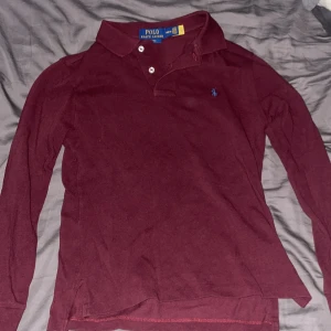 Vinröd pikétröja från Polo Ralph Lauren - Snygg vinröd långärmad pikétröja från Polo Ralph Lauren i slim fit. Klassisk krage med knappar och den ikoniska blå loggan broderad på bröstet. Tillverkad i mjuk bomull som är skön mot huden. Perfekt för dig som gillar stilrena och tidlösa plagg.