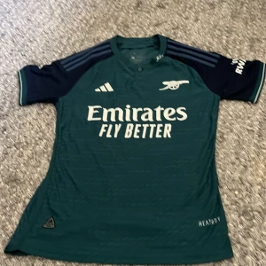 Arsenal bortatröja 23/24 Adidas M - Arsenal bortatröja säsong 23/24 från Adidas i mörkgrönt med marinblå ärmar och vita detaljer. Klassisk kanonlogga på bröstet, broderad Adidas-logga och tryckt sponsor. Tillverkad i lätt funktionsmaterial med subtilt mönster och HeatReady-teknologi.