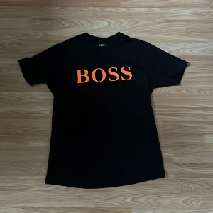Svart BOSS t-shirt med orange tryck - Svart t-shirt från BOSS med stort orange BOSS-tryck på bröstet. Klassisk rund hals och korta ärmar. Tillverkad i 100% bomull för en skön och mjuk känsla. Perfekt för dig som gillar stilrena och tydliga loggor. Lite mindre än L. 
