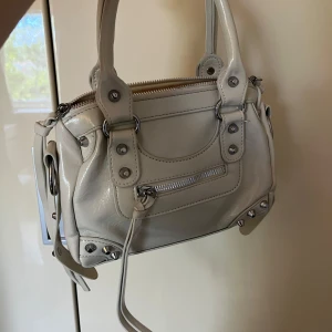 Beige handväska med nitar och dragkedja - Snygg beige handväska i skinn med coola silverfärgade nitar och detaljer. Väskan har både handtag och dragkedja upptill samt en mindre ficka med dragkedja framtill. Perfekt storlek för det viktigaste!