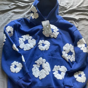 Blå hoodie Denim Tears - Säljer en blå hoodie från Denim Tears med vita bomullsinspirerade tryck över hela tröjan. Tröjan har huva, magficka och ribbade muddar. Perfekt statement-plagg för dig som vill sticka ut!            - Pris är ej hugget i sten! Kom med förslag 💰